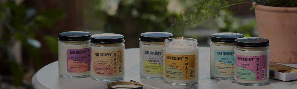 image of candles in clear jars with bright and colorful labels. One candle is lit. In a lifestyle setting with plants in the background