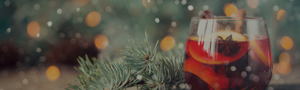 close up of a red holiday punch with clove, start anise, and oranges in a clear stemless wine glass with pine decoration next to it and a depth of field background