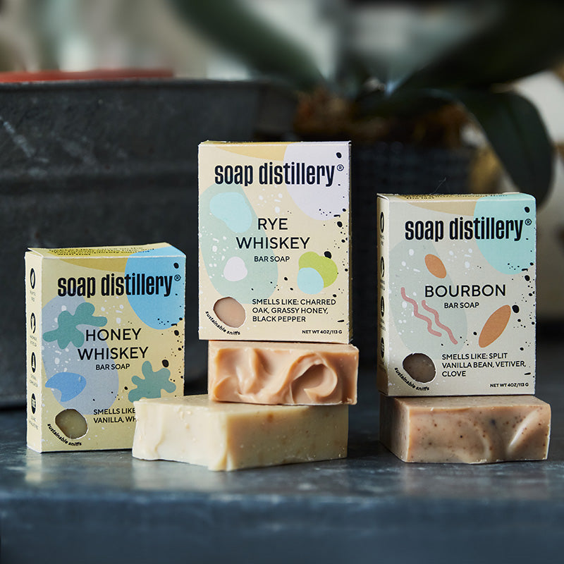 three bar soaps in colorful packaging (honey whiskey, rye whiskey, and bourbon) on a dark grey concrete table with a grey plant pot in the background