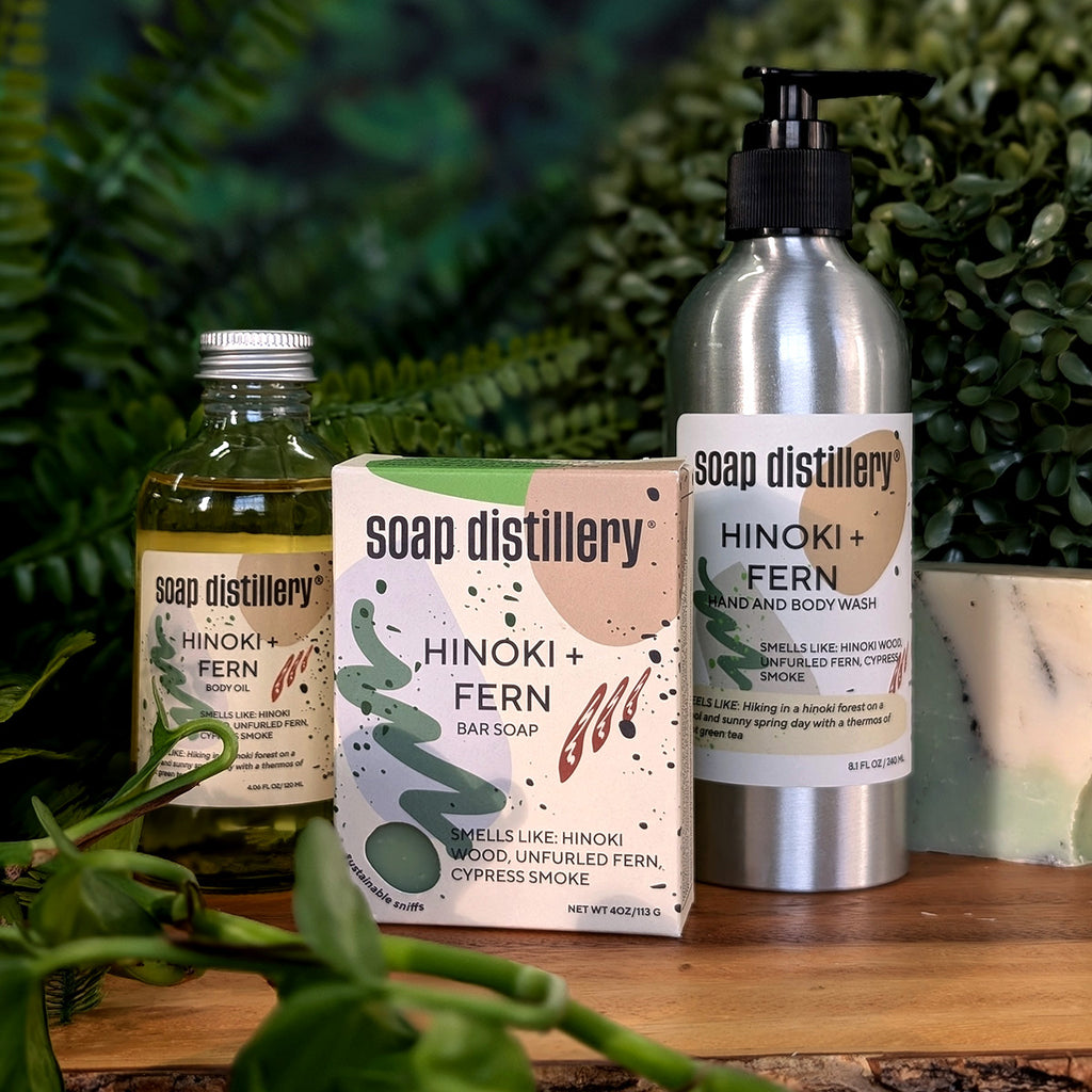 golden body oil in glass bottle with silver cap and tan colored label along with a tan printed box with soap and a silver aluminum body wash bottle on a wooden surface with plants in the background