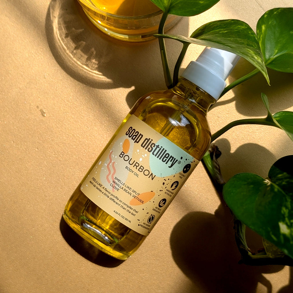 Aerial view of the Bourbon Botanical Body Oil in glass packaging with a white pump top and colorful tan colored label bathed in sunlight with strong shadows
