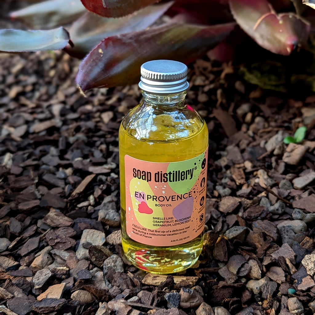 en provence body oil sitting in small landscape gravel near a pink and green bromeliad