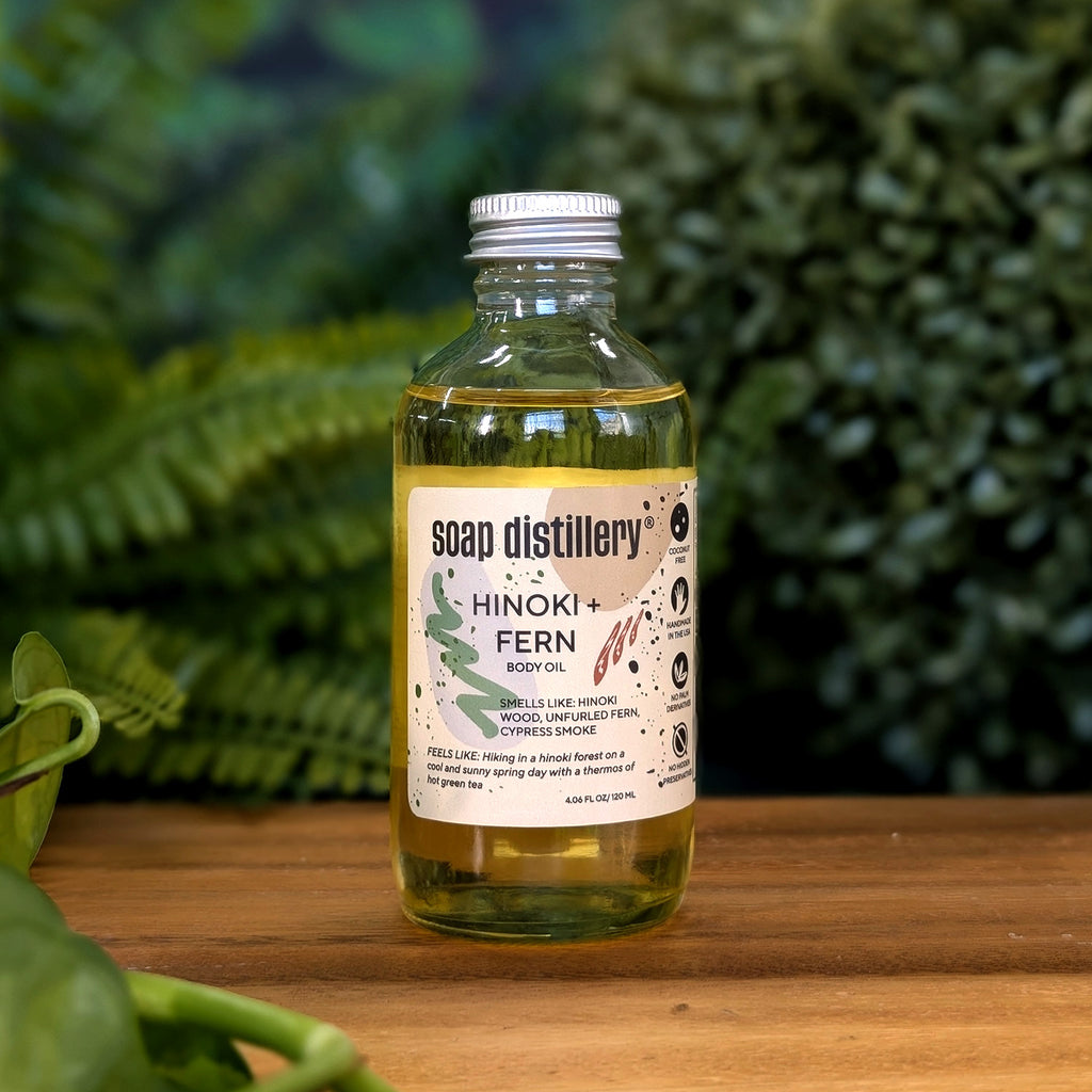 golden body oil in glass bottle with silver cap and tan colored label on a wooden surface with plants in the background