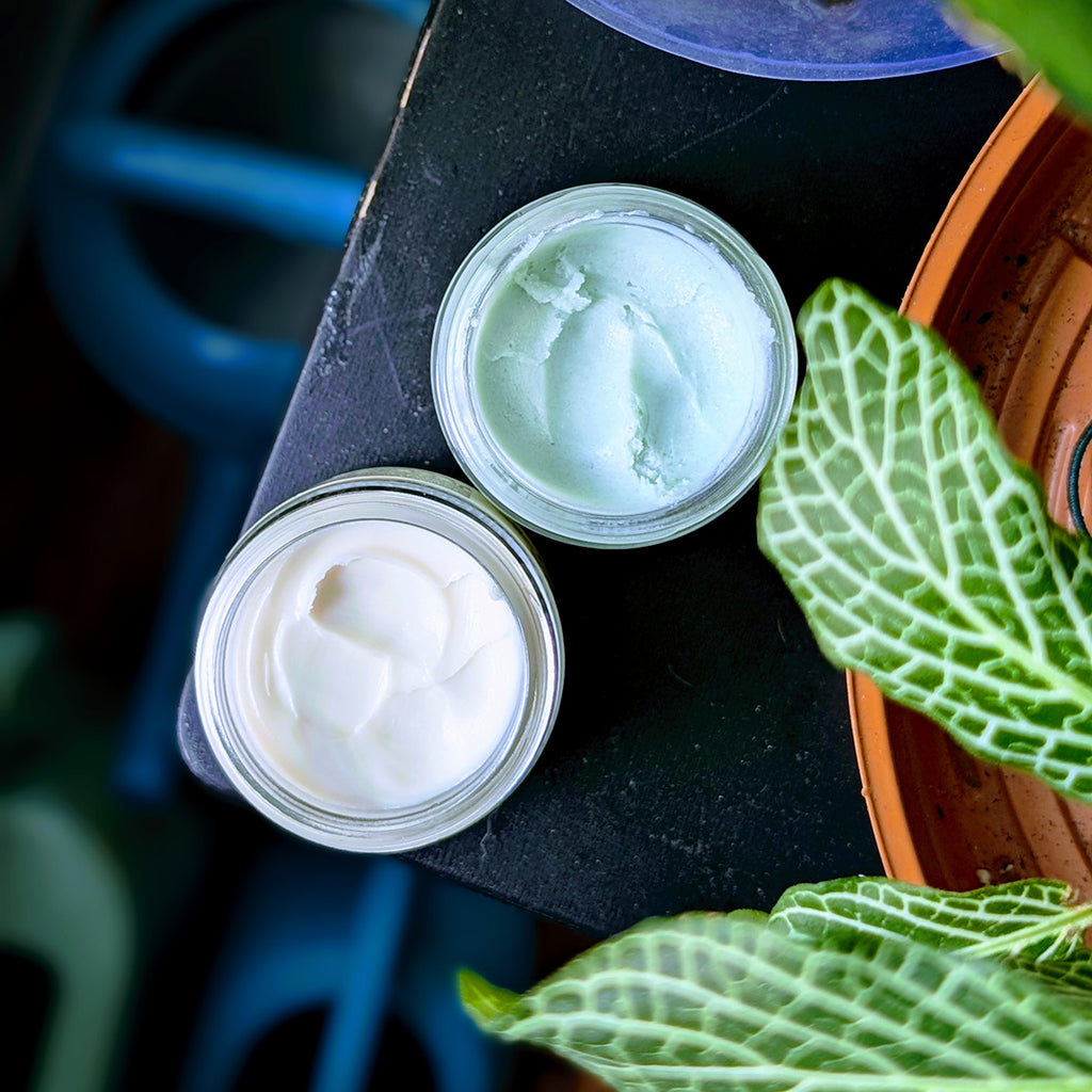 Two jars, one of cream and one of a light green scrub, on a dark surface with green leaves around