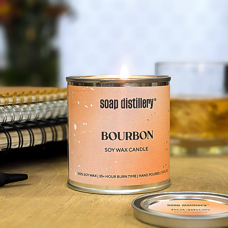 candle in a small paint can with an orange and tan label on a wooden surface with depth of field books and whiskey in the background