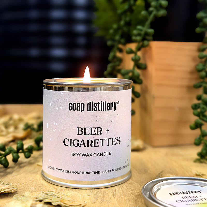 candle in a small paint can with a grey and green label on a wooden surface with depth of field plants