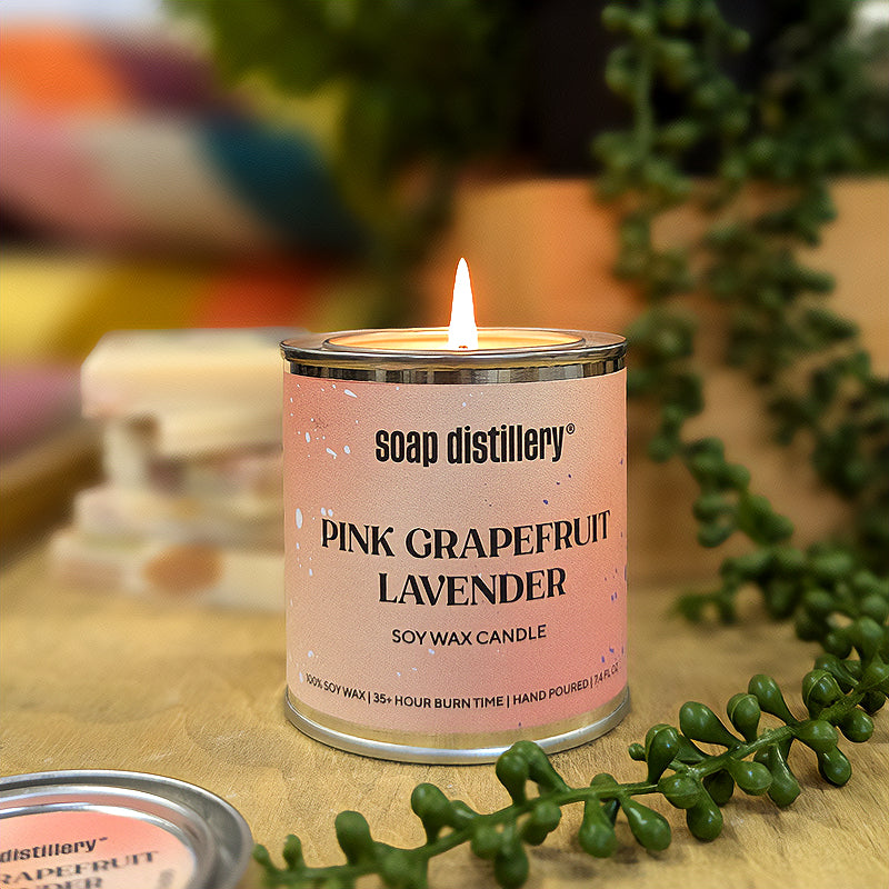 candle in a small paint can with a peachy pink colored label on a wooden surface with depth of field plants and colorful objects in the background