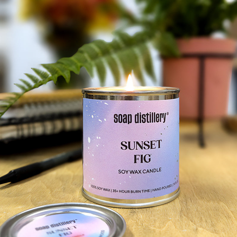 candle in a small paint can with a purple and lavender colored label in a wooden surface with depth of field plants and notebooks in the background