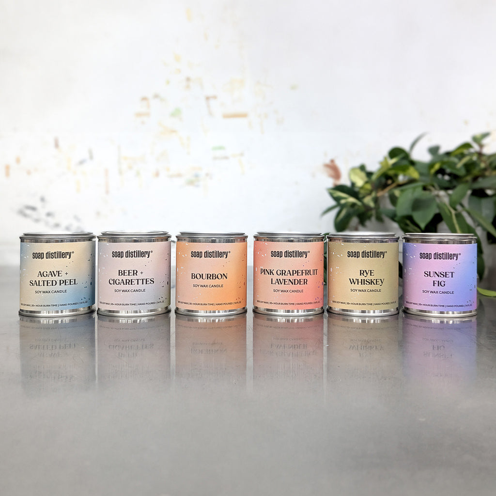 6 candles in small paint cans with colorful labels on a reflective surface