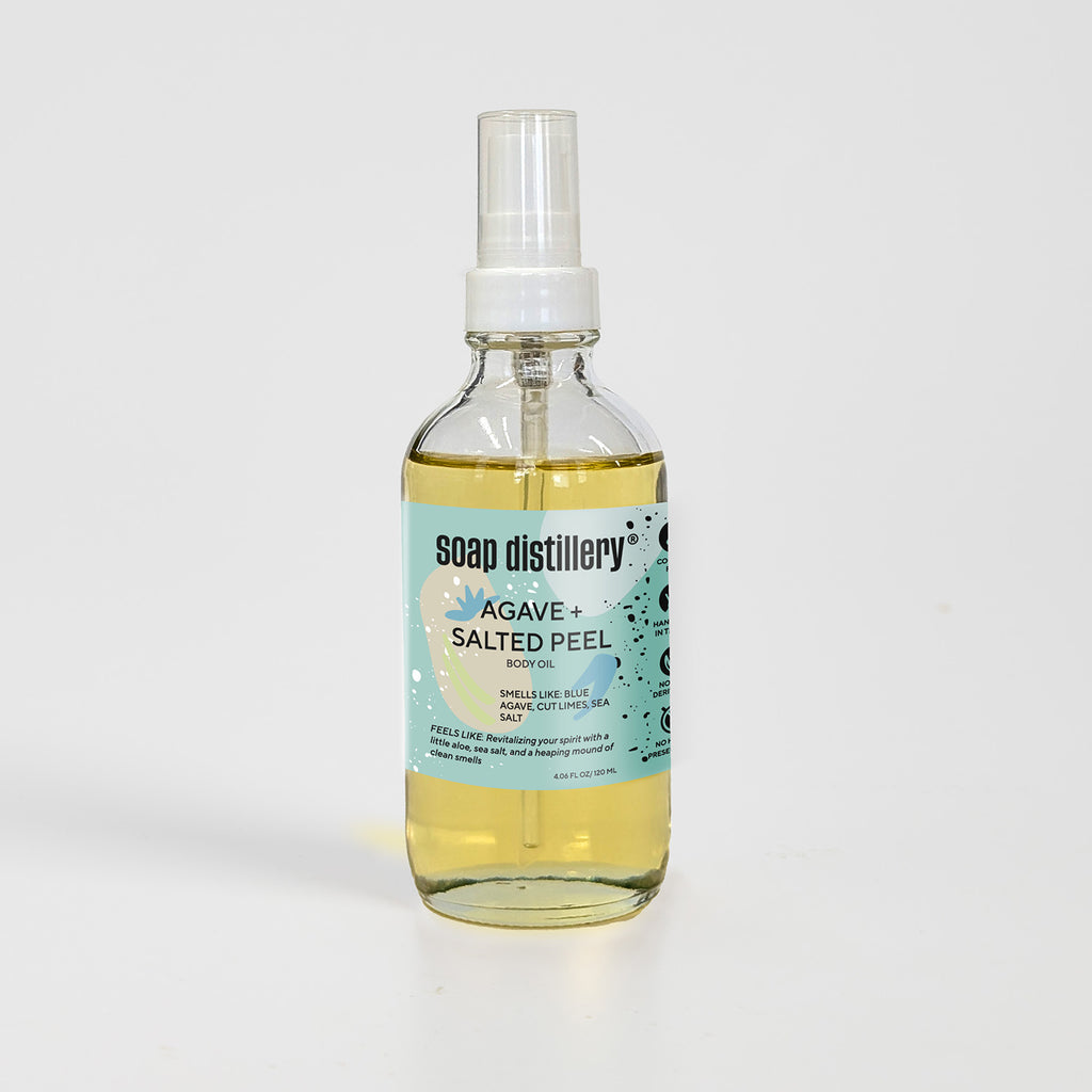 golden body oil in glass bottle with white pump top and seafoam green colored label against a light grey background. Scent is called agave + salted peel