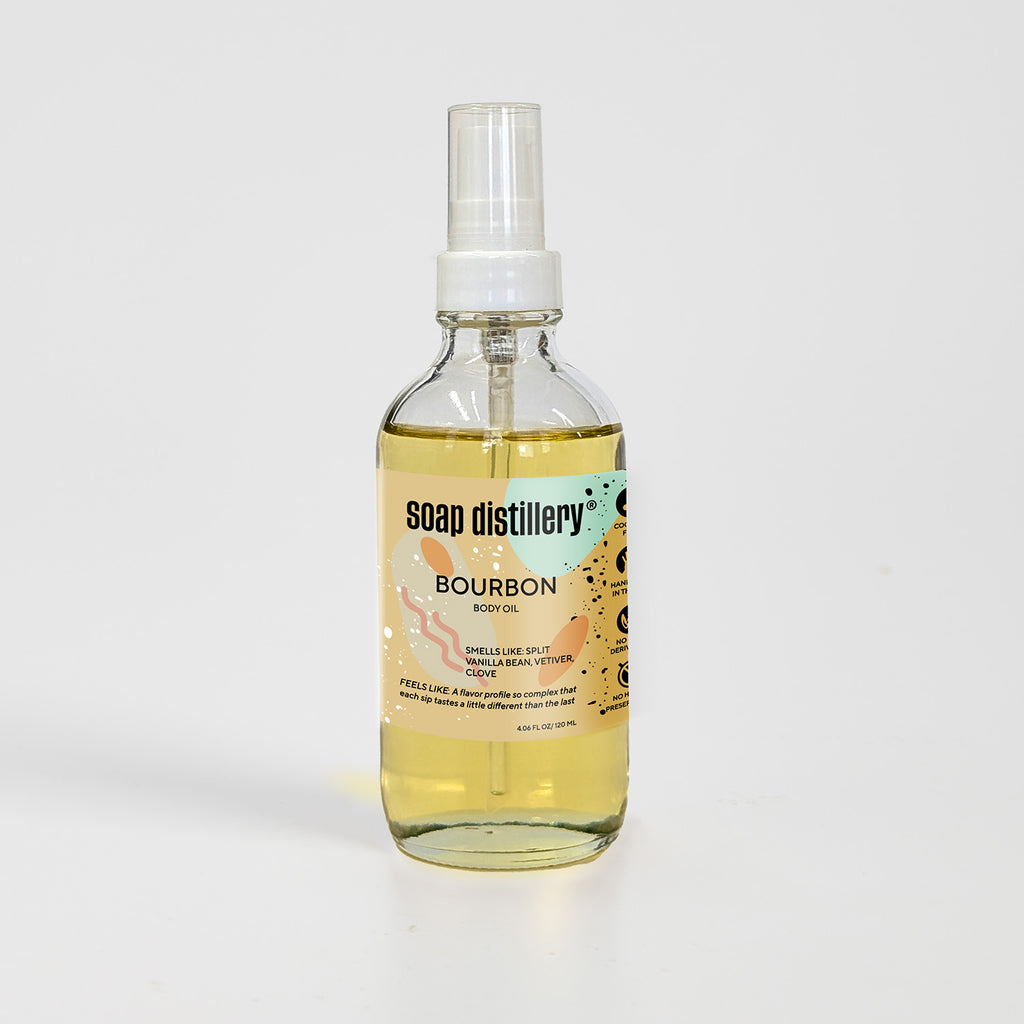 golden body oil in glass bottle with white pump top and light brown colored label against a light grey background. Scent is called bourbon