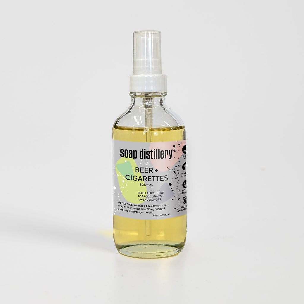 golden body oil in glass bottle with white pump top and light grey colored label against a light grey background. Scent is called beer + cigarettes