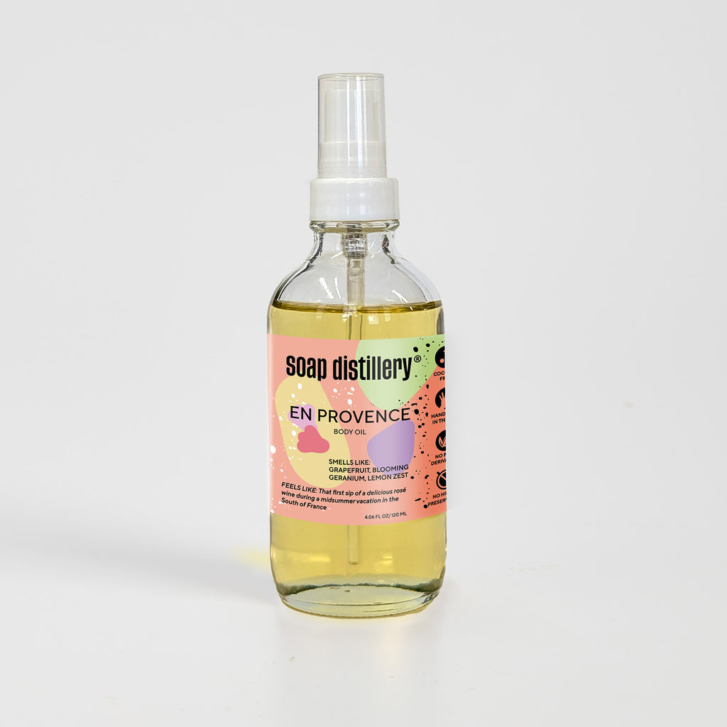 golden body oil in glass bottle with white pump top and coral colored label against a light grey background. Scent is called en provence
