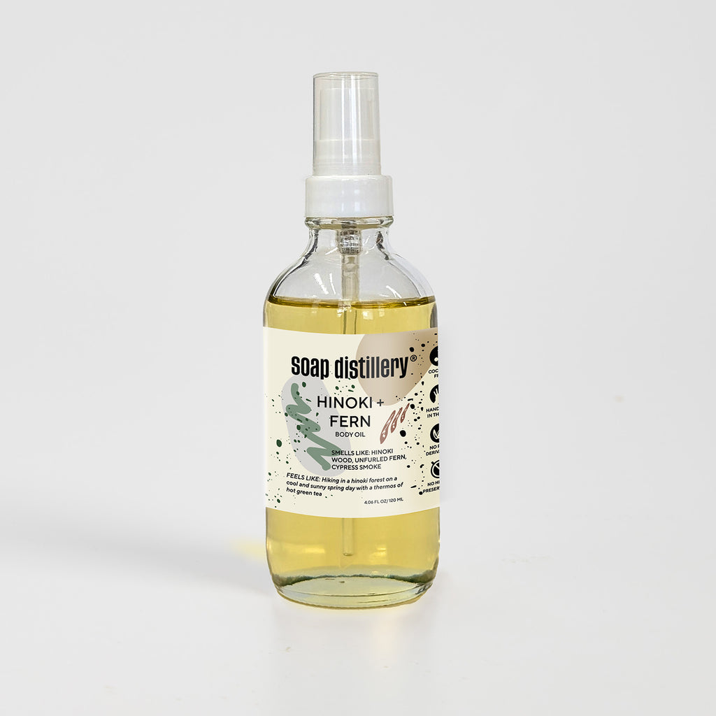 golden body oil in glass bottle with white pump top and tan colored label against a light grey background. Scent is called "hinoki + fern"