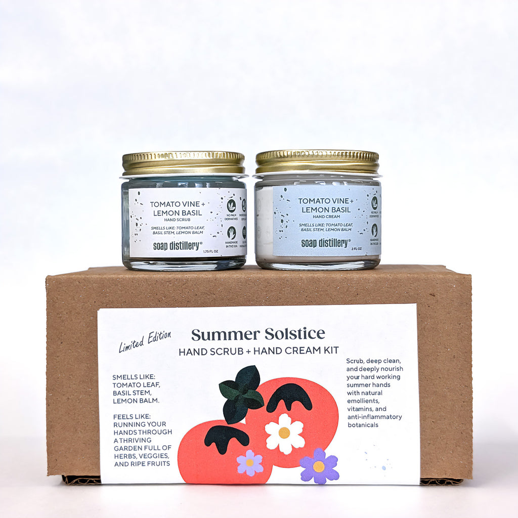 Two jars of soap distillery products (hand scrub + hand cream) on a cardboard box with 'Summer Solstice' name. Limited edition summer herbs inspired hand care kit.