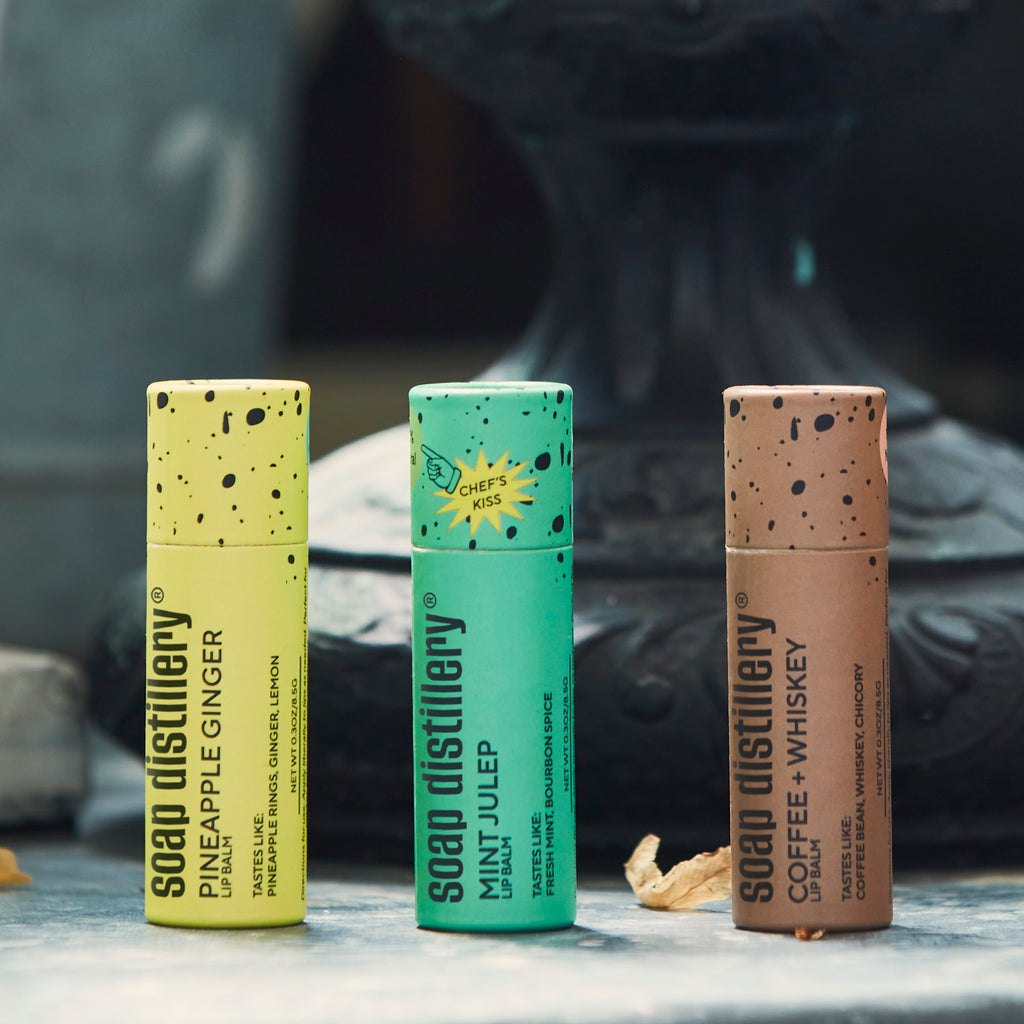 Pineapple ginger, mint julep, and coffee + whiskey lip balms standing on a concrete surface
